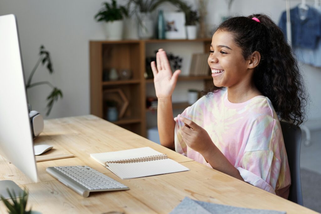 pexels-photo-4143791-4143791 A happy girl waves during an online class at home, sitting at a desk with a computer.